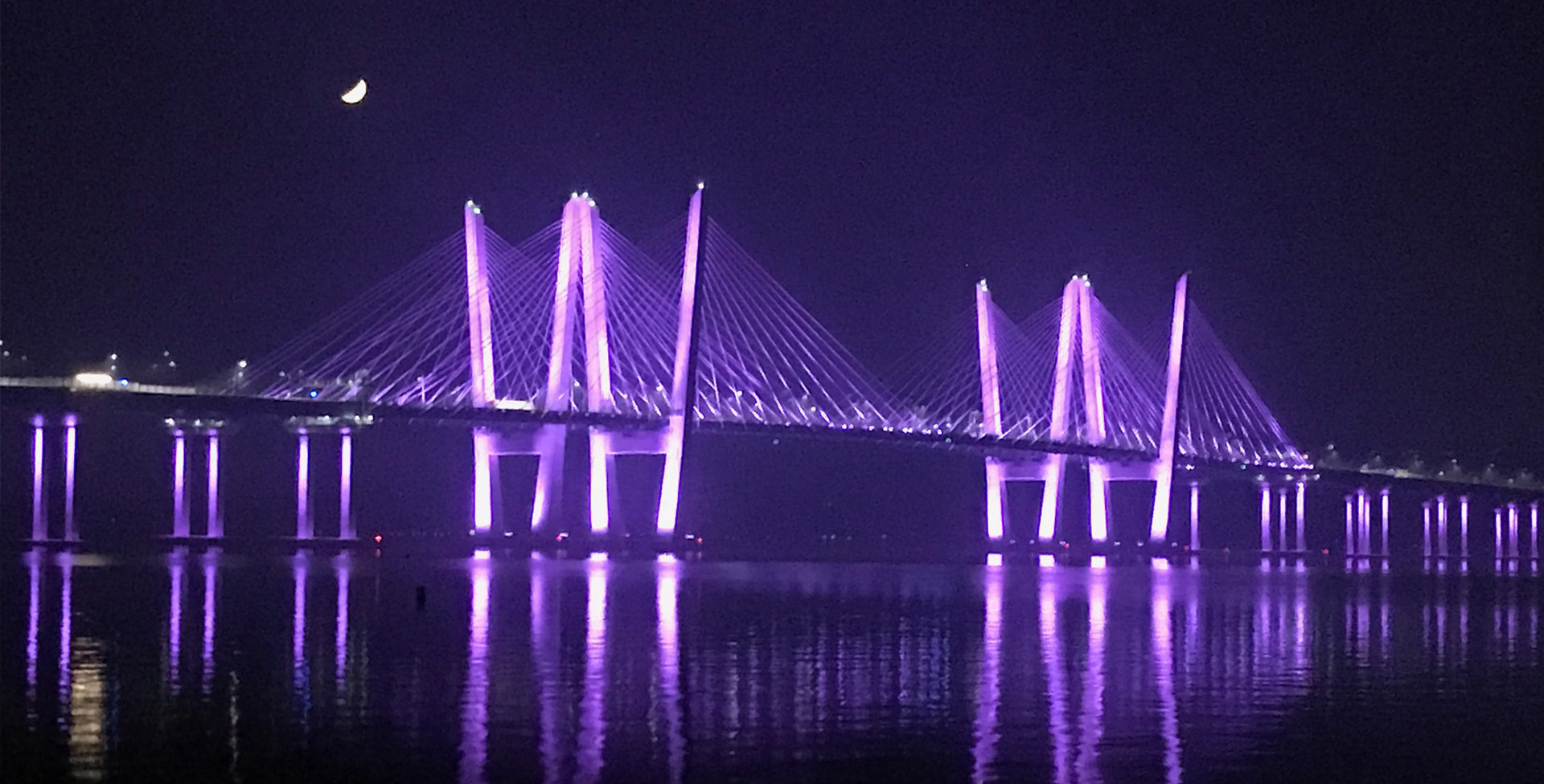 The image displays a nighttime scene of a bridge illuminated with purple lights, spanning over a body of water with reflections visible on its surface.